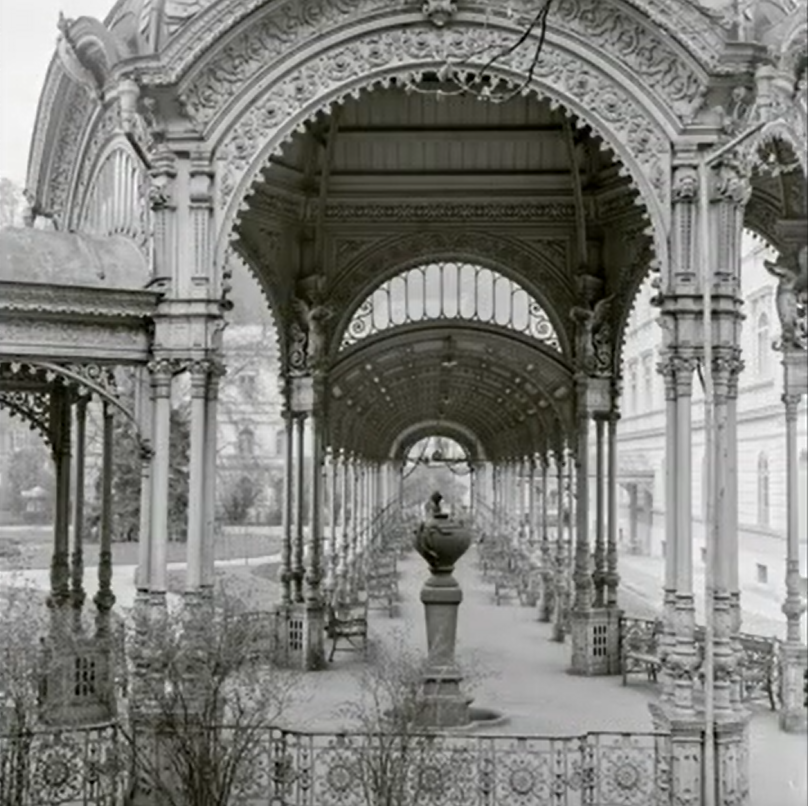 Black and white photograph of an ornate corridor with arches and decorative railings, featuring a central fountain and empty benches along the sides.